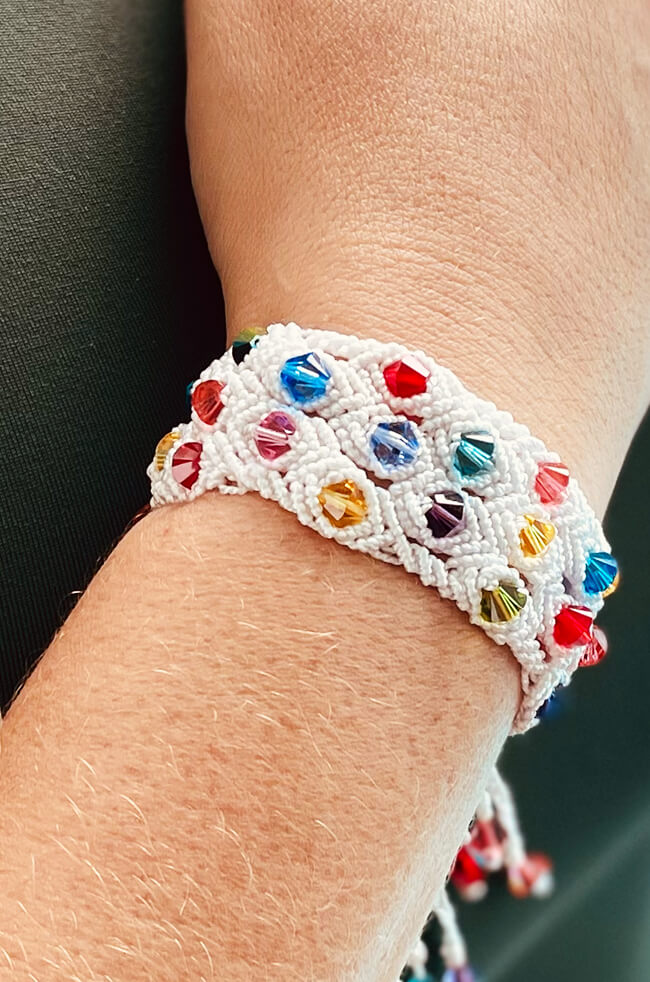 Rainbow in White: A handmade white macramé bracelet adorned with colorful faceted beads in red, blue, yellow, purple, and green, worn on a person's wrist against a dark background.