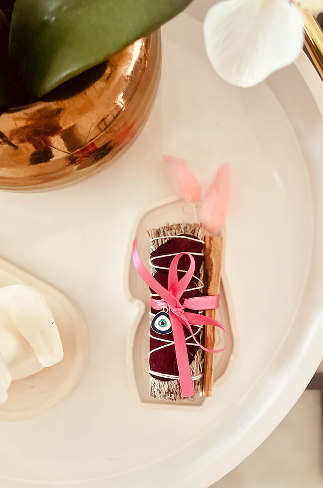 Healing Energy Enhancer Rose Oval Evil Eye Charm Kit: Rose, sage, and Palo Santo bundle tied with a pink ribbon and adorned with an Evil Eye charm, displayed on a white tray.