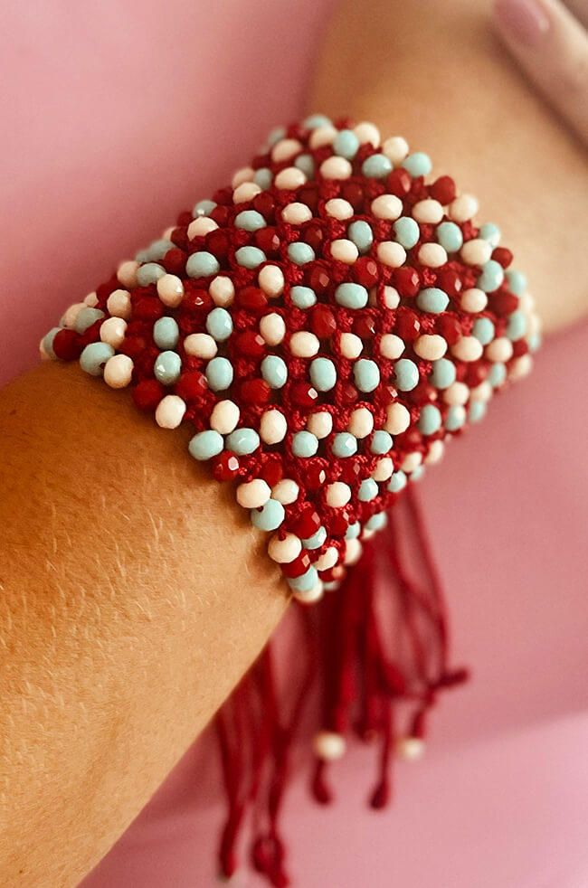 Scarlet: A wide handmade bracelet crafted with red thread and adorned with alternating light blue, white, and red beads, featuring tassel ends that hang loosely.