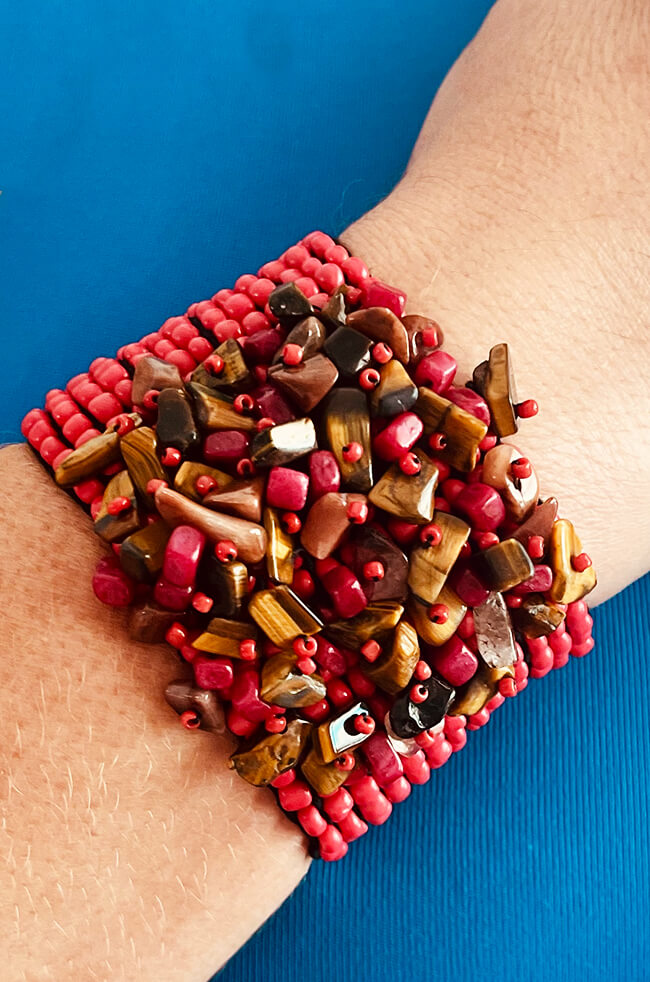 Siena: A bold handmade bracelet featuring clusters of red, brown, and golden tiger's eye chip beads, all strung together over a base of small red seed beads, worn on a wrist against a blue background.