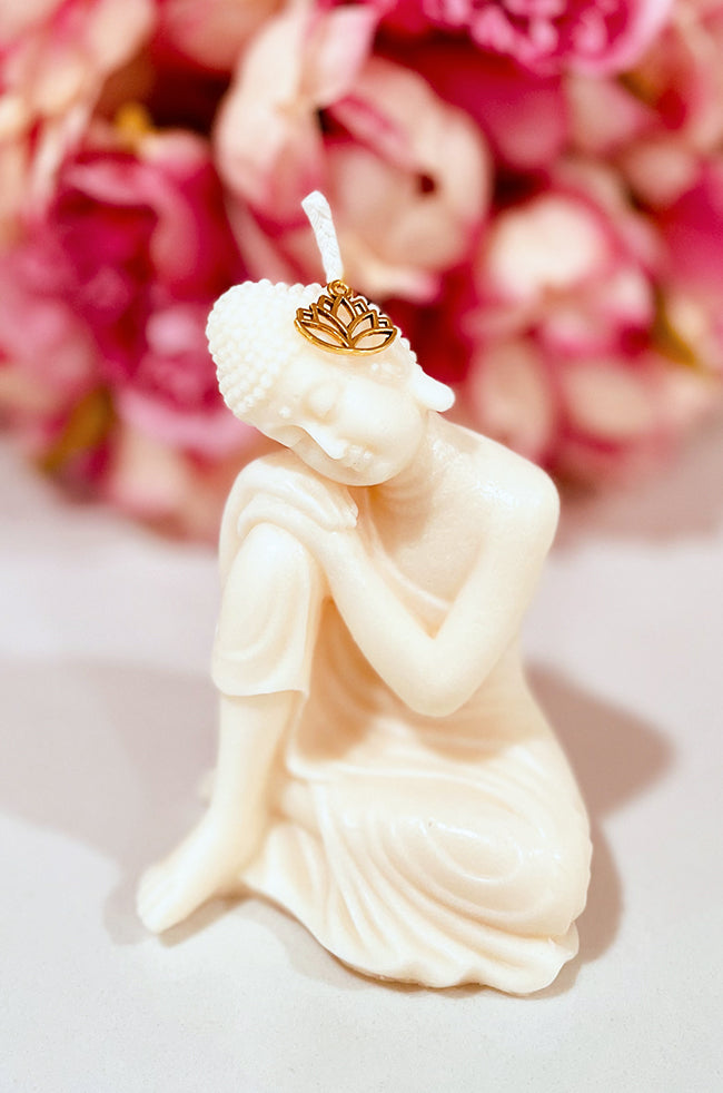 Hand-poured Sleeping Buddha candle with intricate details, resting peacefully with a gold lotus charm on its head, set against a background of pink flowers.