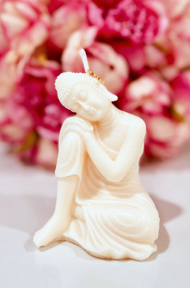 Hand-poured Sleeping Buddha candle with intricate details, resting peacefully with a gold lotus charm on its head, set against a background of pink flowers.