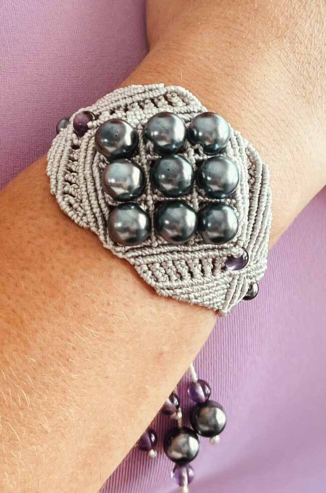 Smoke: A handmade macramé bracelet featuring a woven gray design with nine shiny black beads arranged in a grid pattern, accented with small purple beads, worn on a person's wrist against a lavender background.