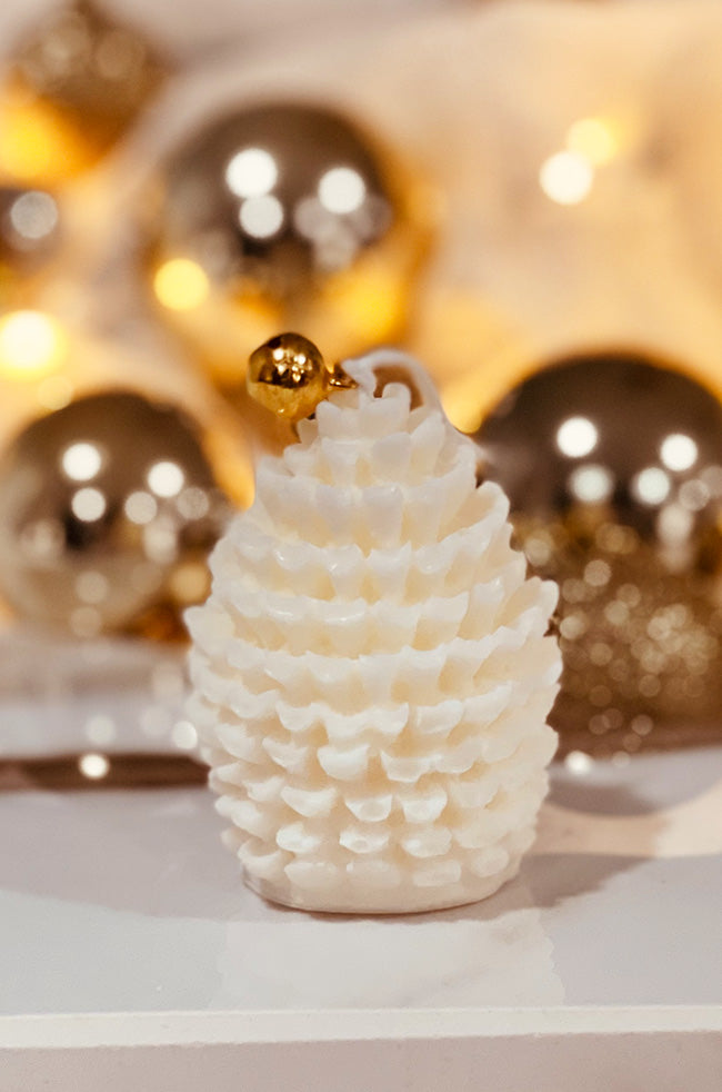 Spicy's Blazing Pine Cone Candle: Ivory pine cone sculptural Christmas candle with layered wax details and a gold bell on top, styled with warm gold holiday ornaments.