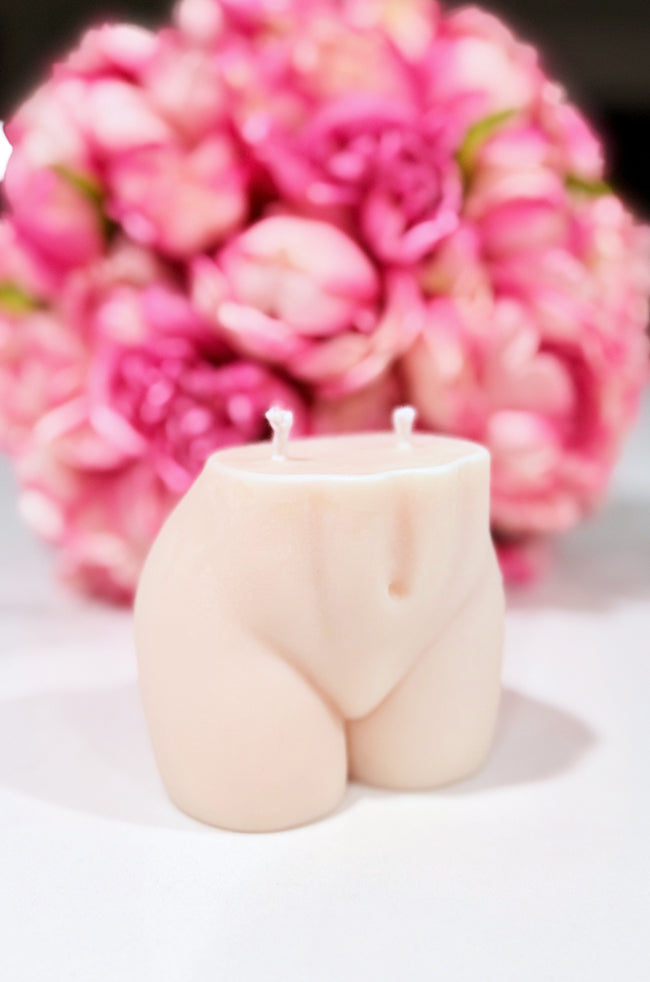 Sultry Candle: A sculpted candle in the shape of a woman's lower torso with dual wicks, set against a soft-focus background of vibrant pink flowers.