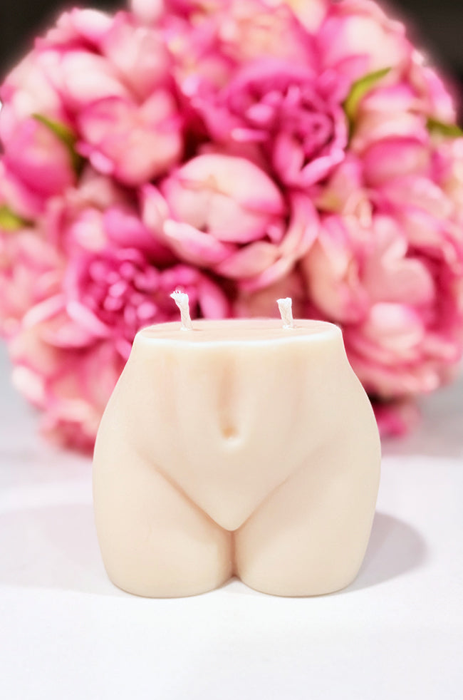 Sultry Candle: A sculpted candle in the shape of a woman's lower torso with dual wicks, set against a soft-focus background of vibrant pink flowers.