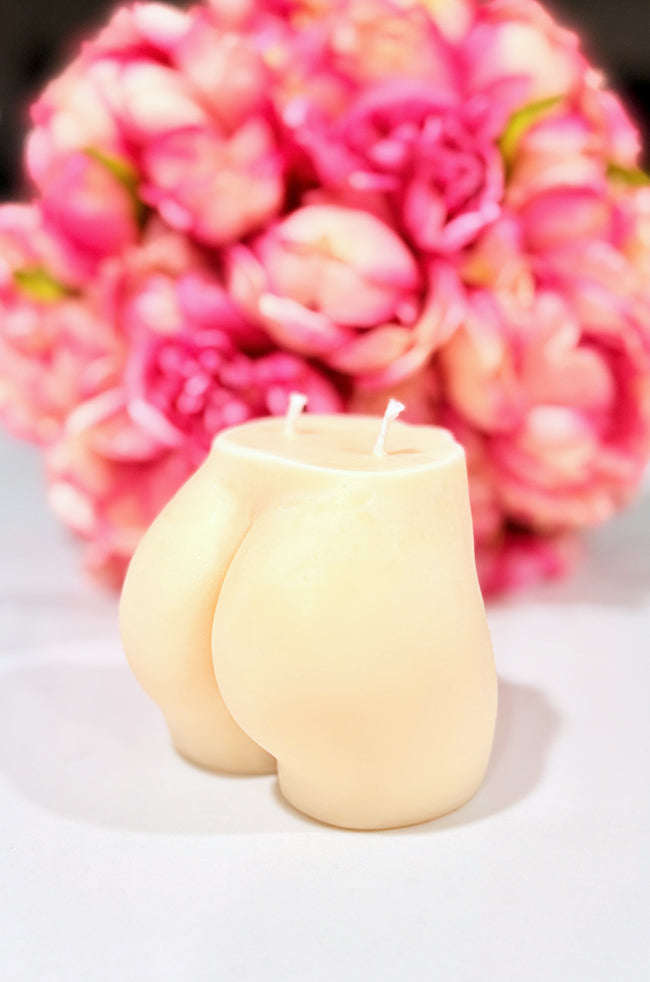 Sultry Candle: A sculpted candle in the shape of a woman's lower torso with dual wicks, set against a soft-focus background of vibrant pink flowers.