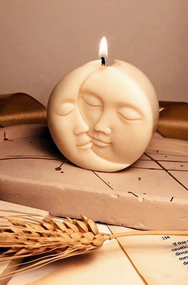 Sun Moon Rising Candle: Lit Sun Moon Rising candle featuring overlapping sun and moon faces, displayed with warm neutral décor and dried wheat.