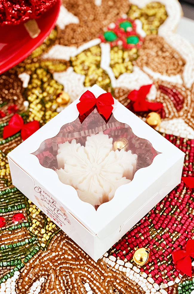 The Christmas Snowflake Candle: Christmas Snowflake sculptural candle displayed in a white gift box with red bow and festive beaded background.