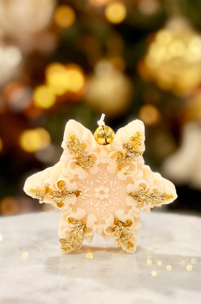 The Renaissance Snowflake Candle: Front view of the ivory Renaissance Snowflake Christmas candle with gold flakes and a gold bell against festive holiday décor.