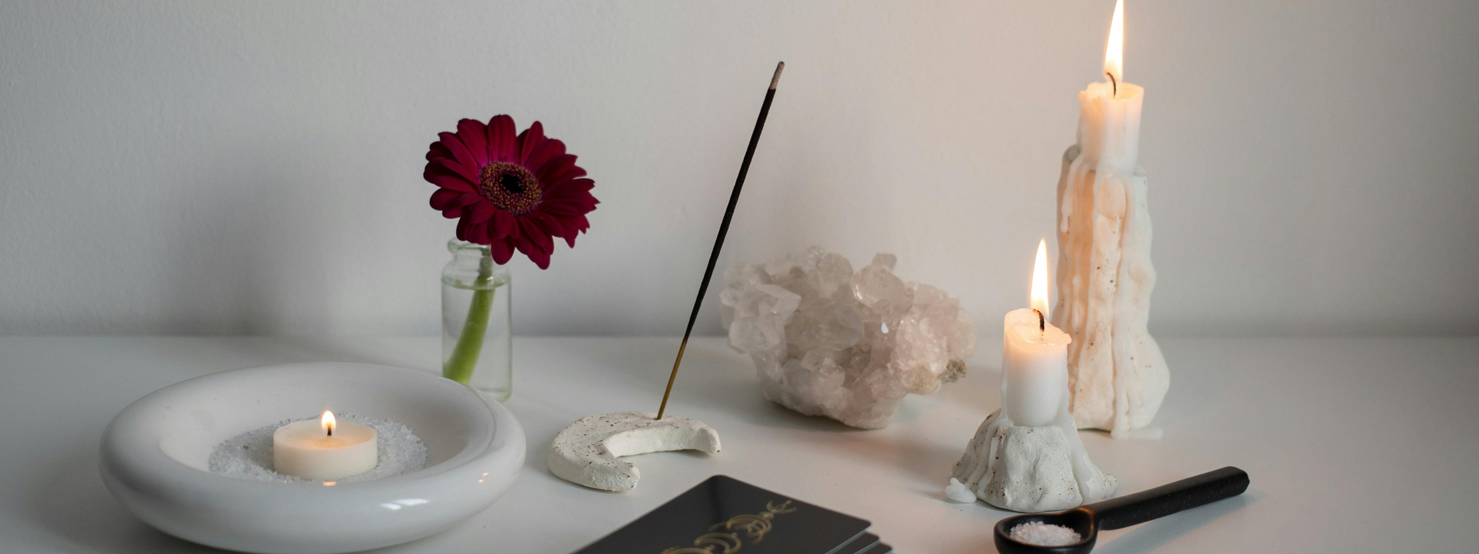 A calming altar setup with lit sculptural candles, a burning incense stick, a red daisy in a glass vase, a white crystal cluster, and a dish of salt on a minimalist white surface.