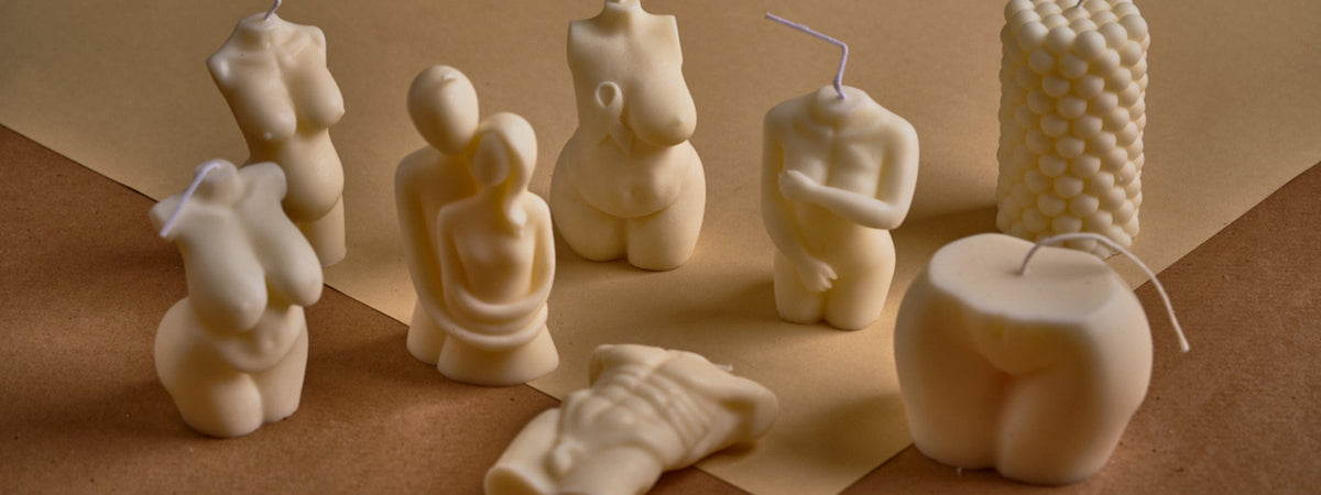 A collection of sculpted cream-colored candles in various artistic shapes, including human torsos, a couple embracing, and geometric designs, displayed on a neutral-toned surface.