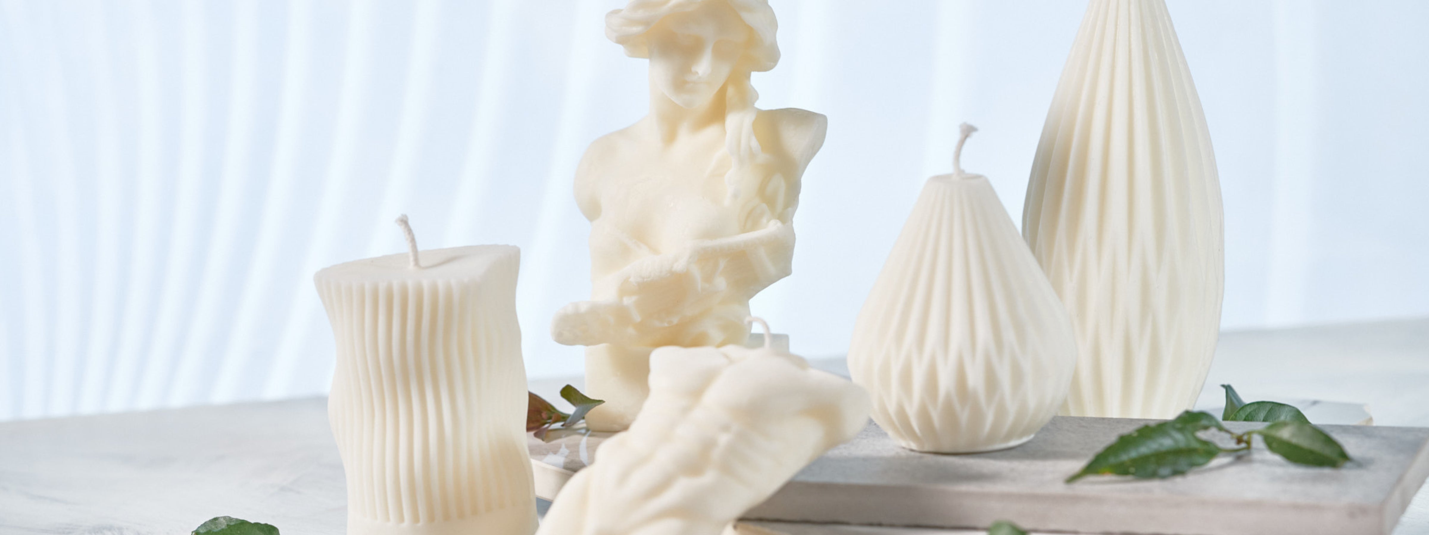 Ivory handmade sculptural candles in geometric and figurative shapes styled with greenery on a white marble surface.