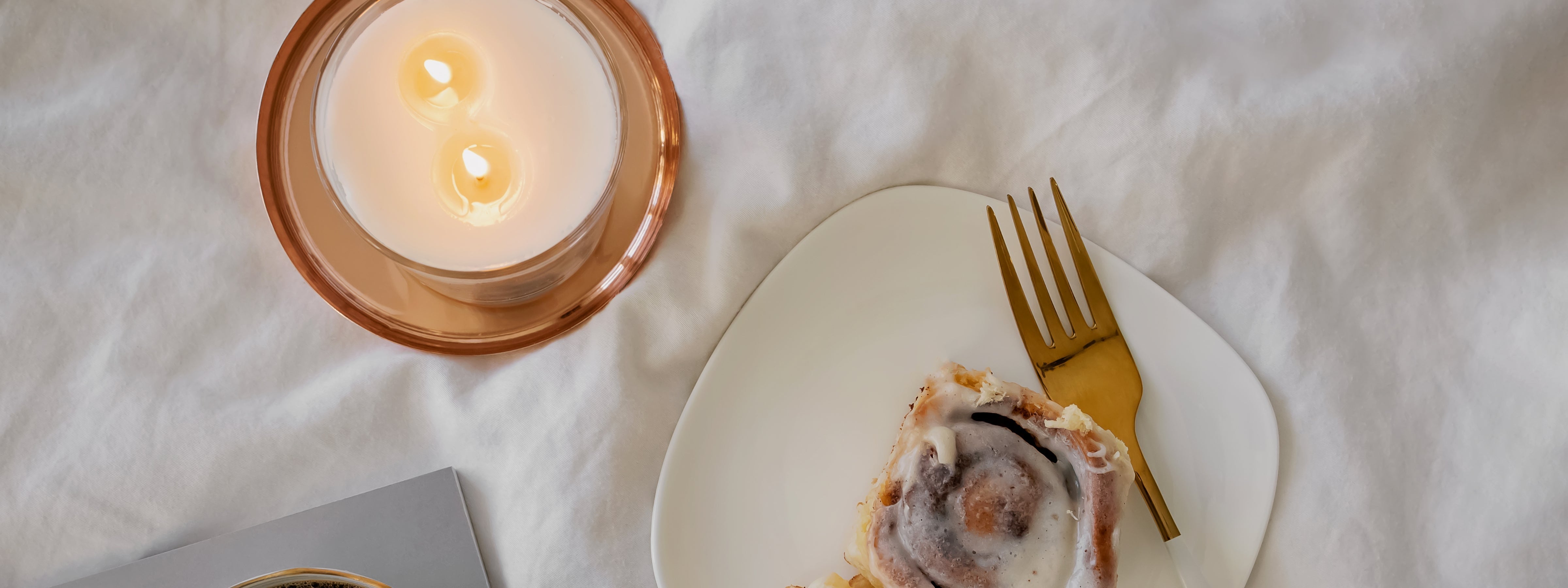 Overhead view of a lit two-wick candle beside a white plate holding a cinnamon roll with icing and a gold fork, styled on soft white fabric to evoke a cozy, gourmand-inspired atmosphere.