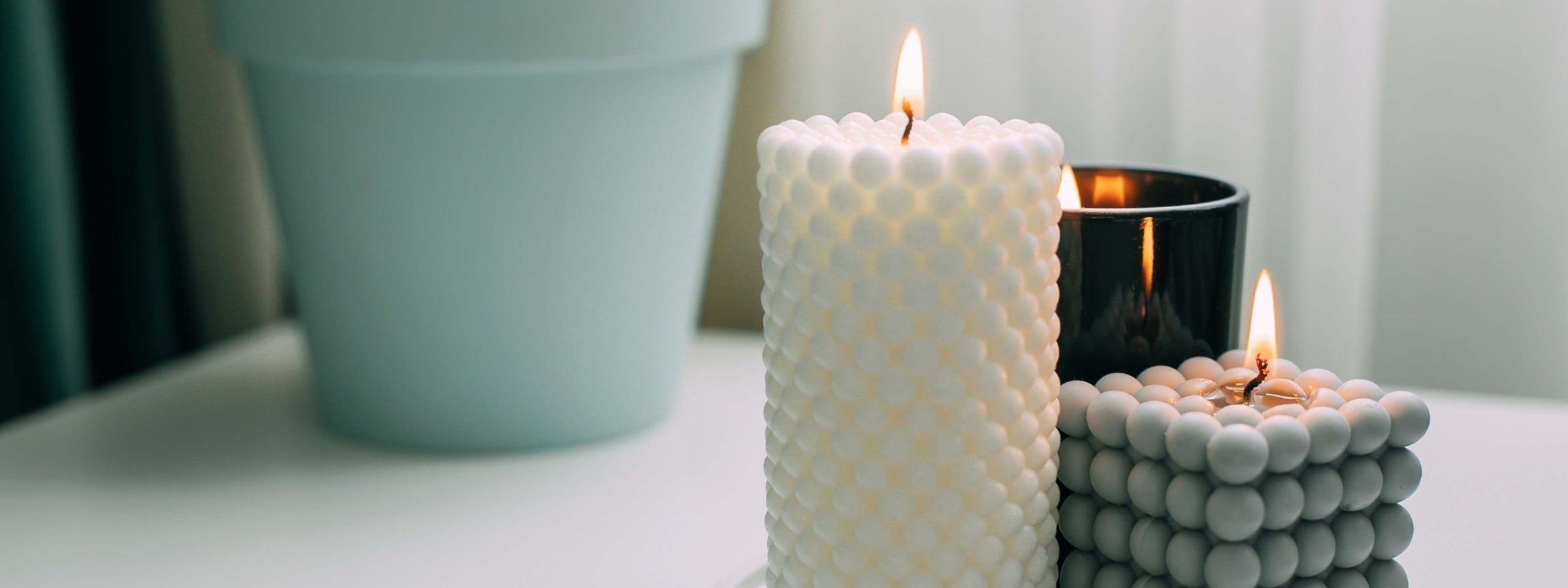 Three lit candles on a white surface: a tall white textured pillar candle, a smaller gray bubble cube candle, and a black glass jar candle, with a blurred light blue pot in the background.