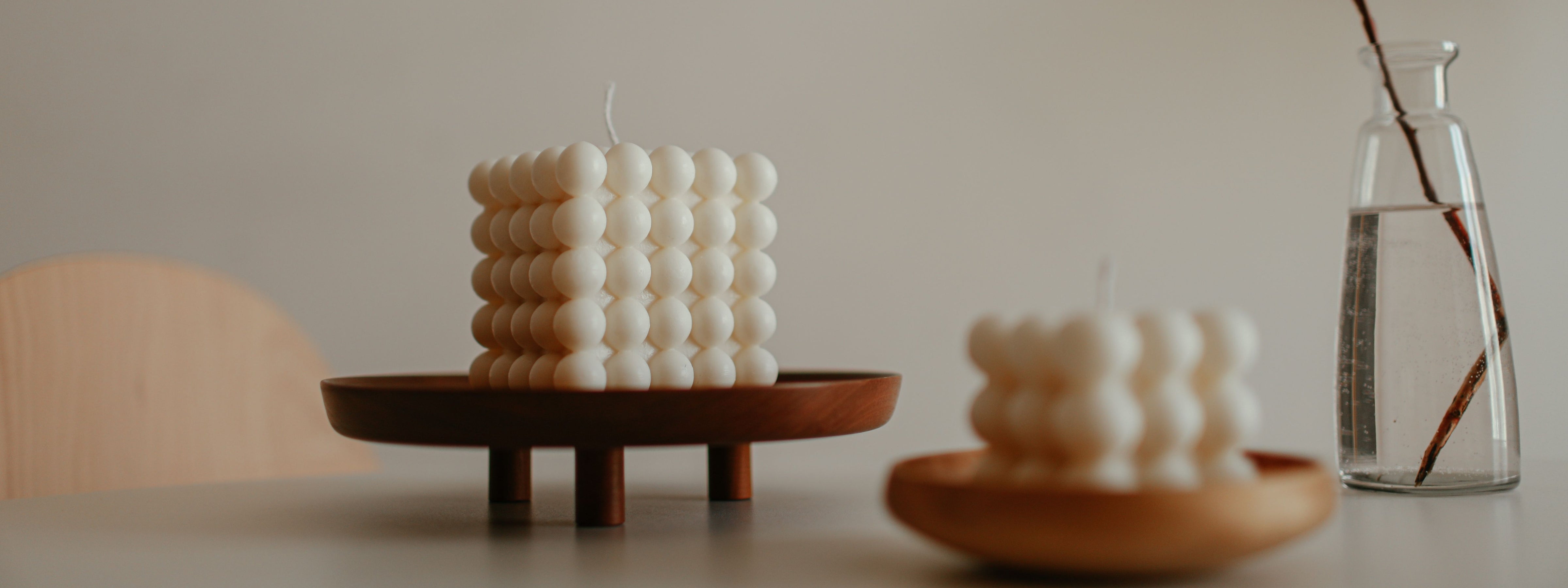 Two white bubble sculptural candles displayed on wooden trays beside a glass vase in a warm, minimalist room.