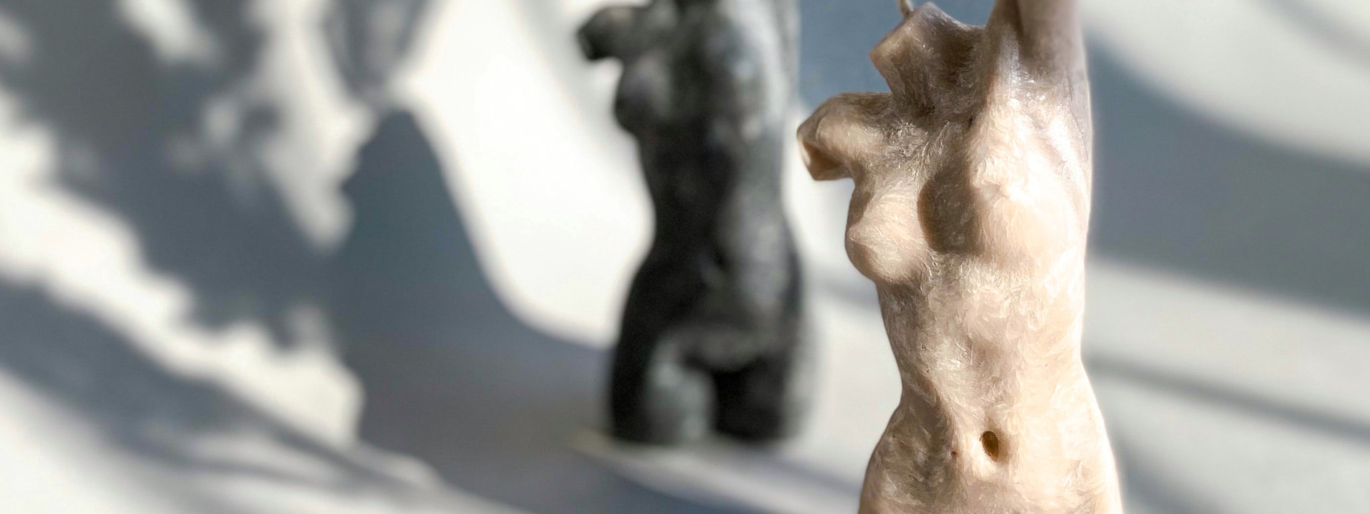 Close-up of a sculptural torso candle in natural sunlight with soft shadows and other decorative candles blurred in the background.