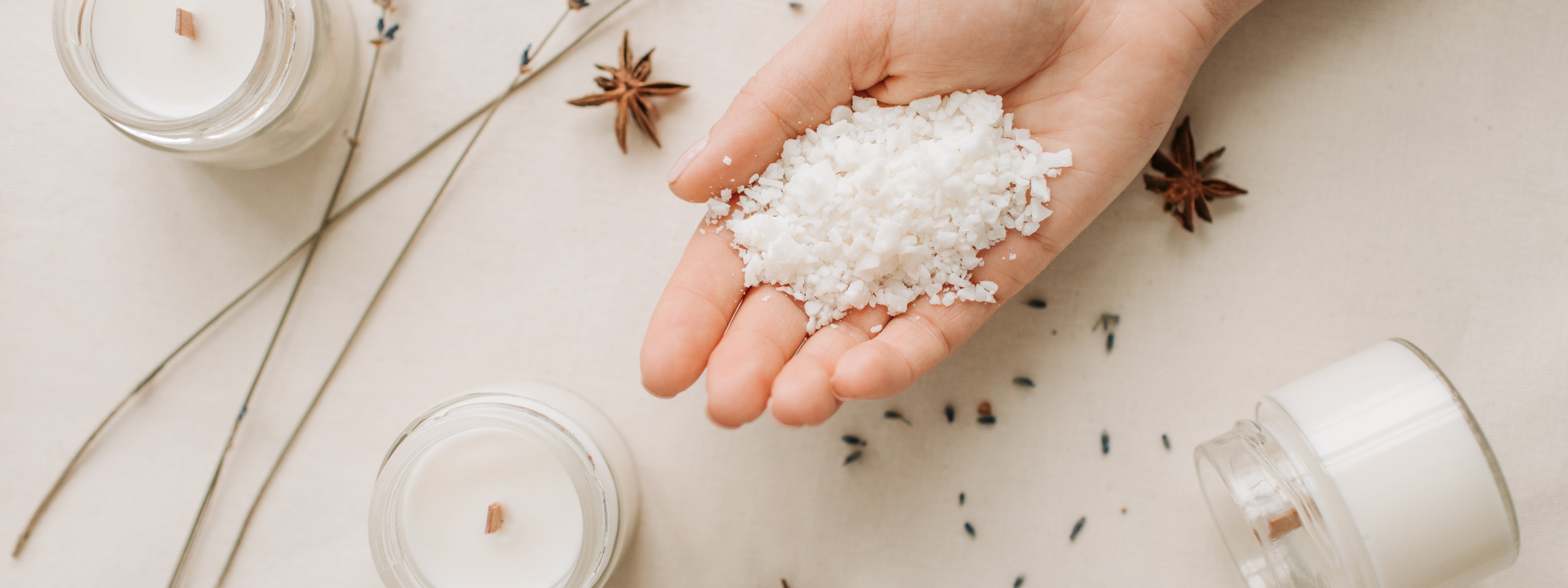 A hand holding natural soy wax flakes beside glass jars of white hand-poured candles with wooden wicks, surrounded by dried star anise and lavender on a neutral linen surface.