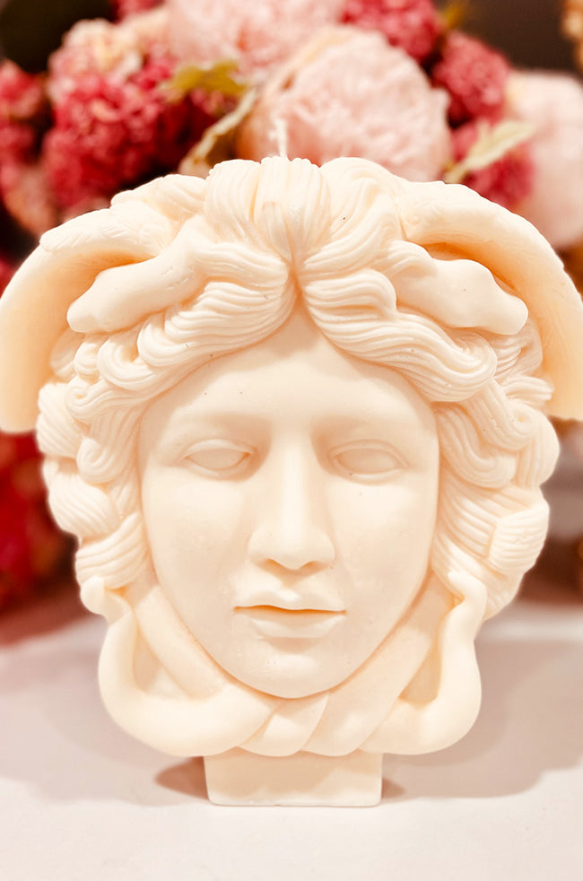 Medusa Candle: Ivory sculpted candle shaped like Medusa's head, featuring intricate serpent hair and classical facial details, styled with soft pink florals in the background.