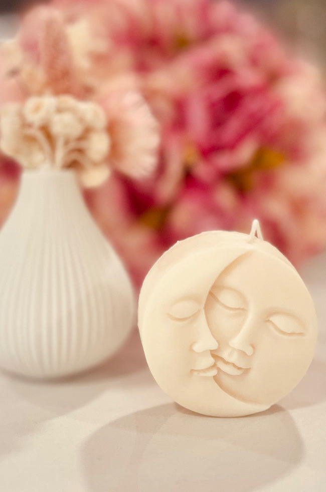 Sun Moon Rising Candle: Ivory Sun Moon Rising sculptural candle with intertwined celestial faces, styled with soft pink flowers in the background.