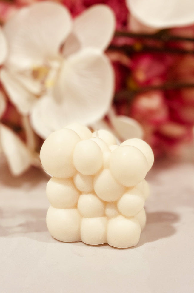 Bubble Cloud: A creamy white bubble cloud candle with an organic, clustered sphere design, displayed on a smooth surface with soft white and pink flowers blurred in the background.