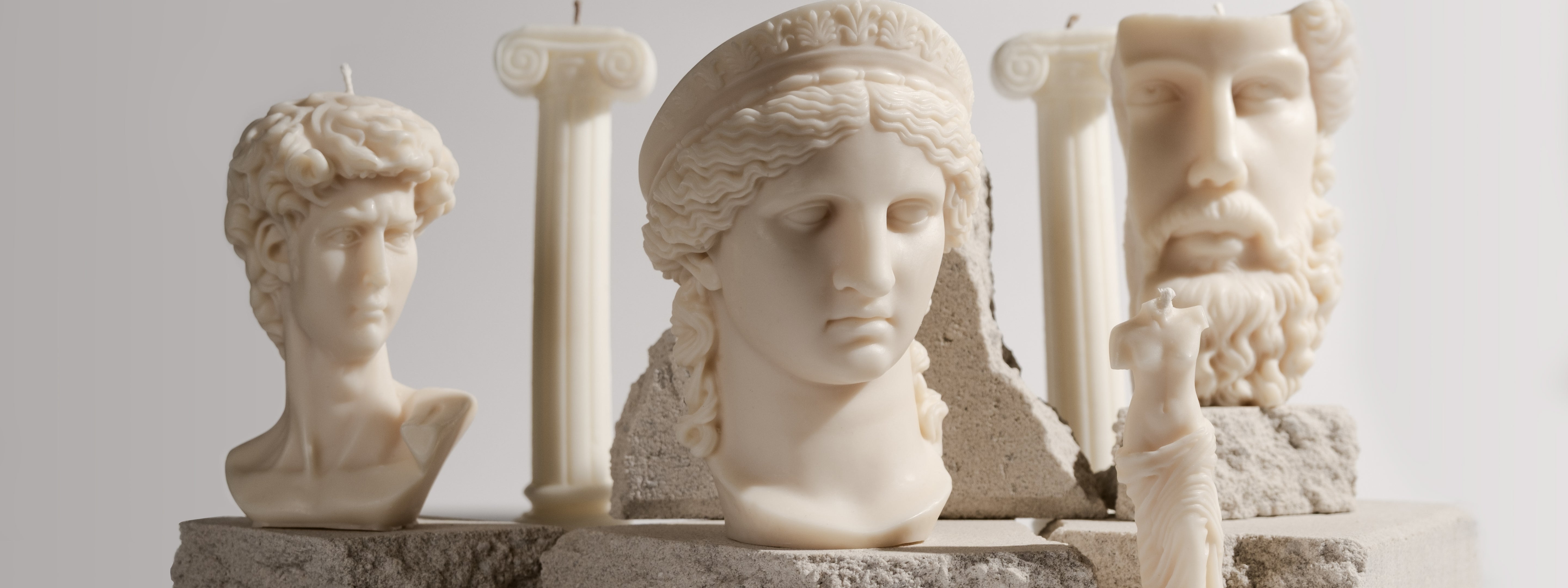A collection of ivory-toned sculptural candles inspired by classical Greco-Roman statues and columns, arranged on stone-textured blocks for a minimalist, artistic display.