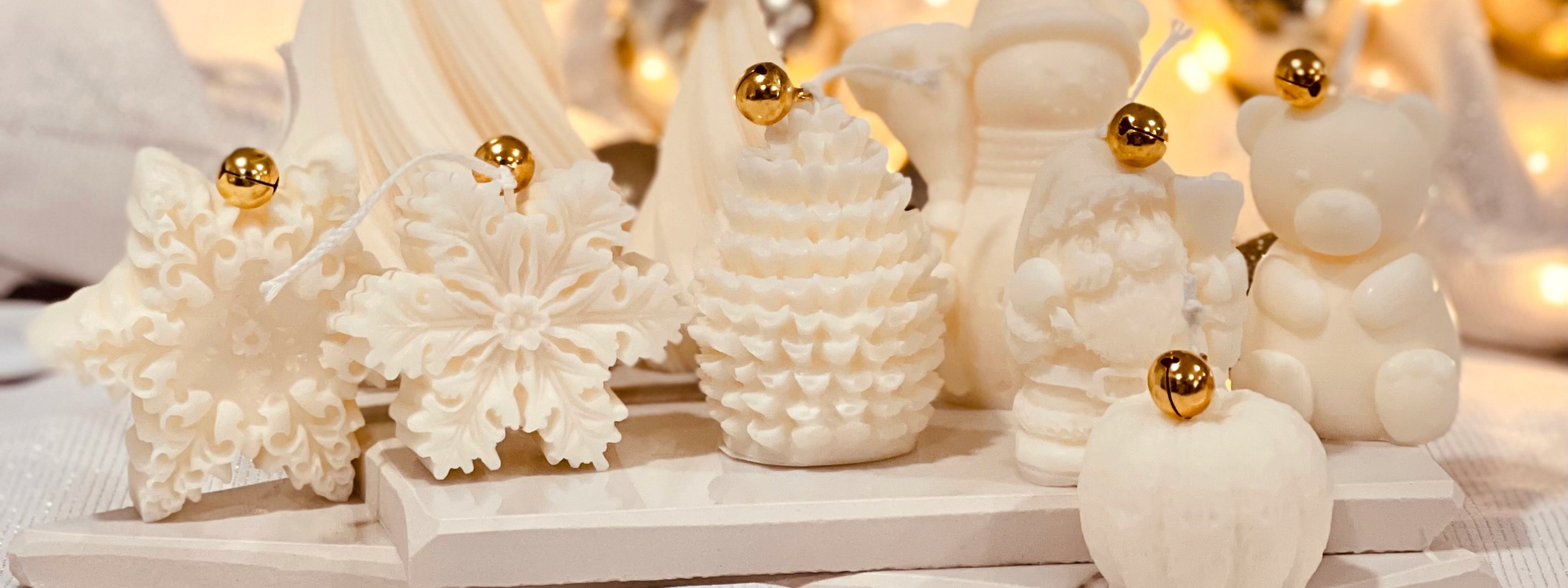 Christmas Collection Candles: A festive display of handmade white sculpted candles shaped like Christmas trees, snowflakes, Santa Claus, and a teddy bear, each topped with a small gold bell. The candles are arranged on a soft white cloth with warm twinkling lights and gold ornaments in the background, creating a cozy holiday atmosphere.