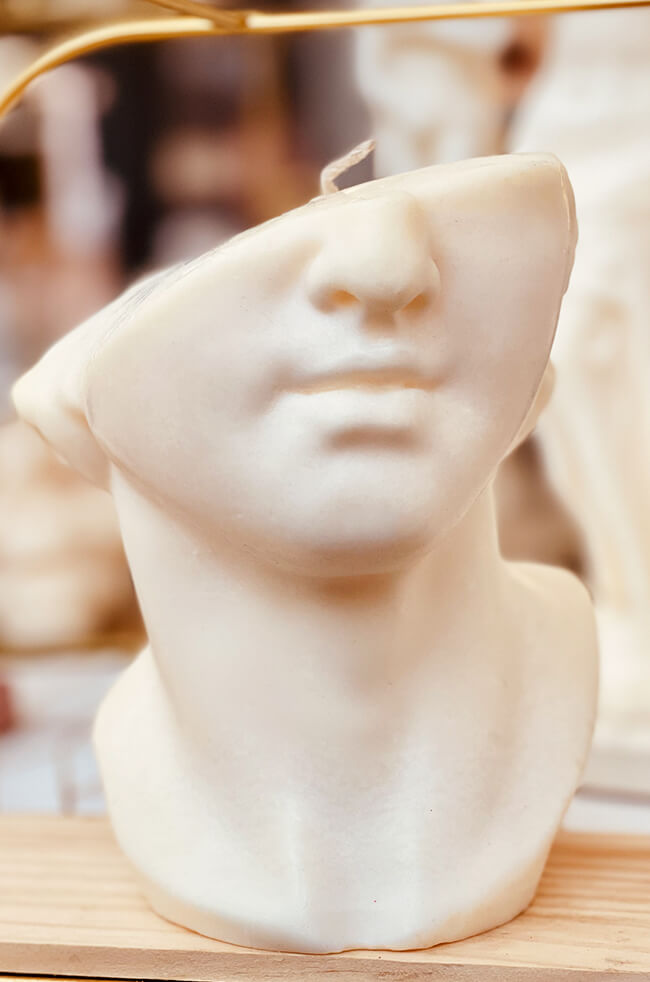 David's Flame: Sculpted white candle resembling a classical male bust with a partially abstract face, displayed on a wooden tray.