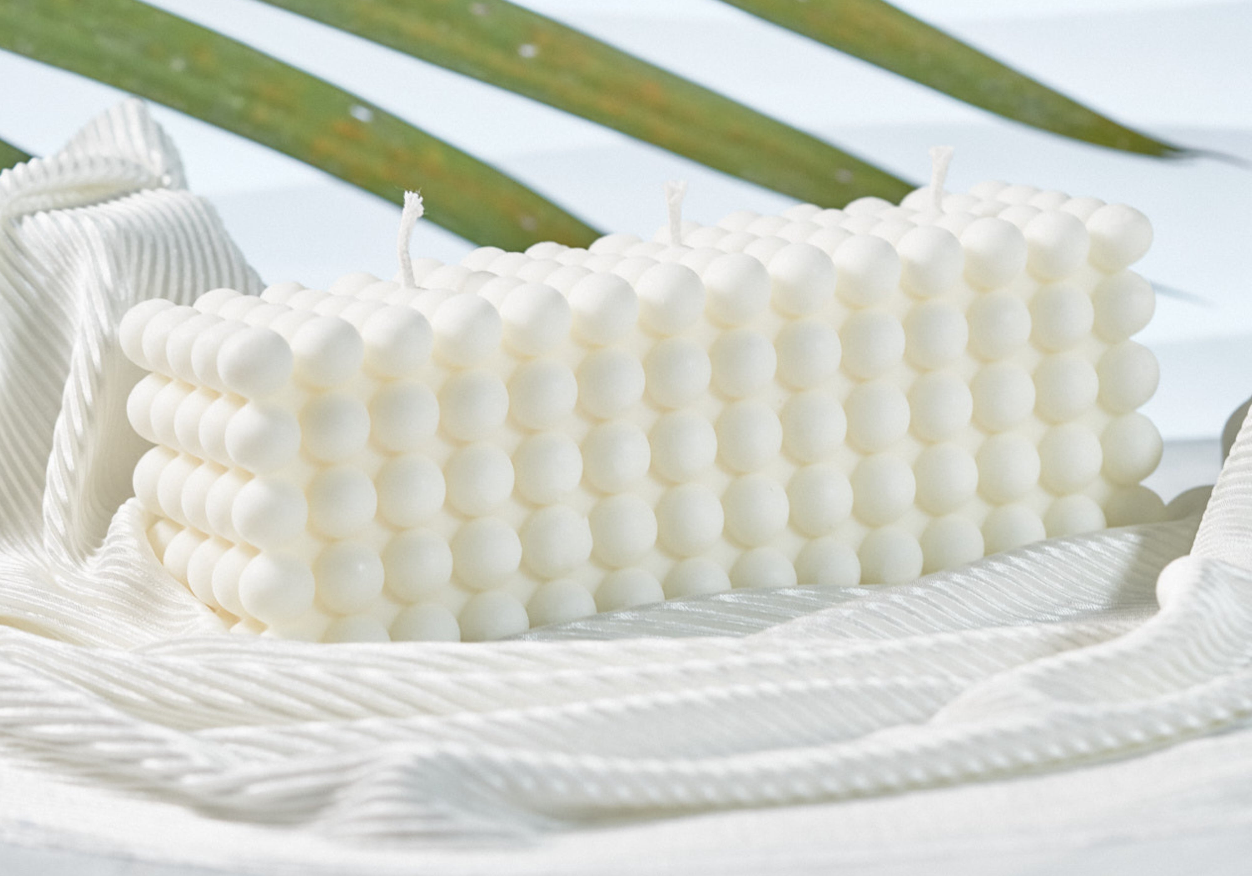 A rectangular white candle with a textured, beaded bubble pattern, resting on a white woven textile with green palm leaves in the background.