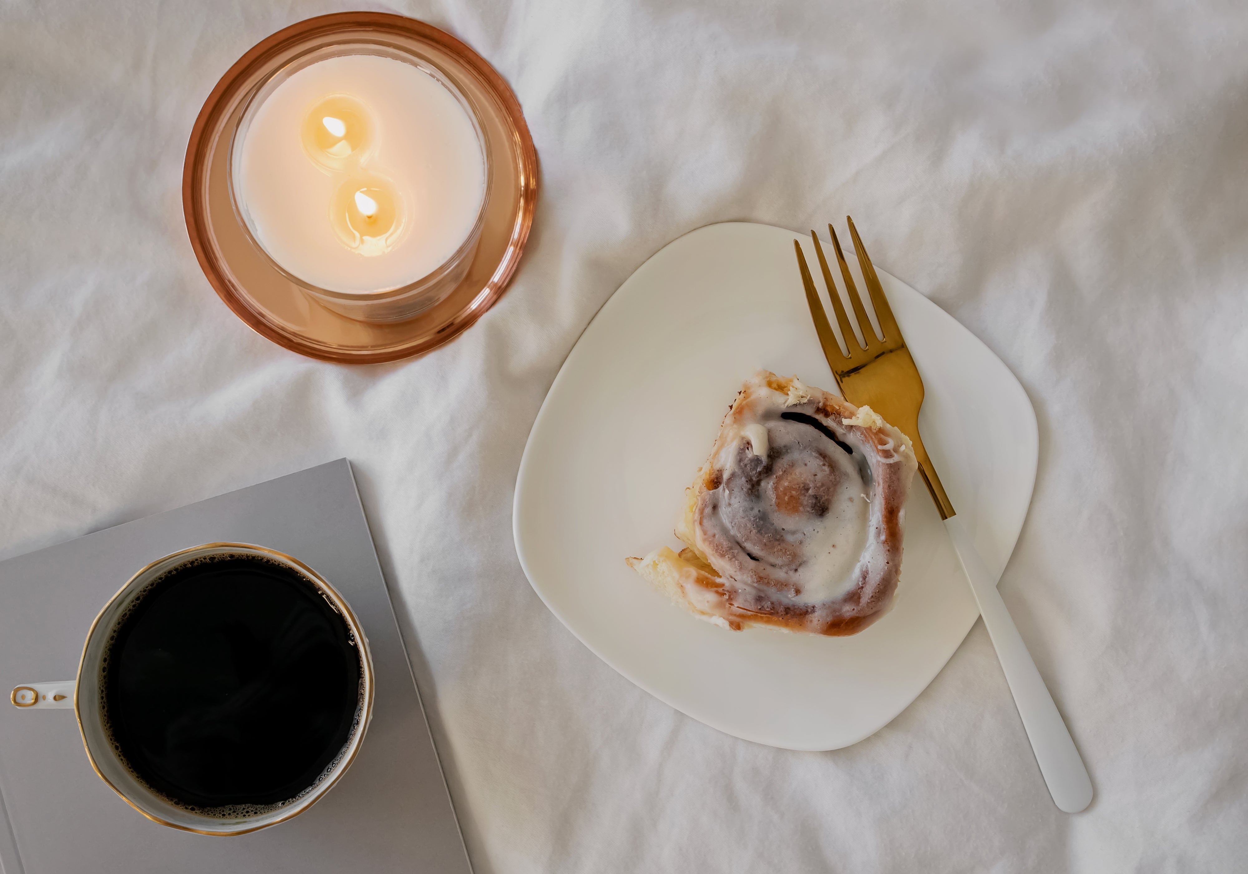 A styled flat-lay featuring a cinnamon roll on a white plate with a gold fork, next to a cup of black coffee and a lit candle, all arranged on a white textured fabric background.