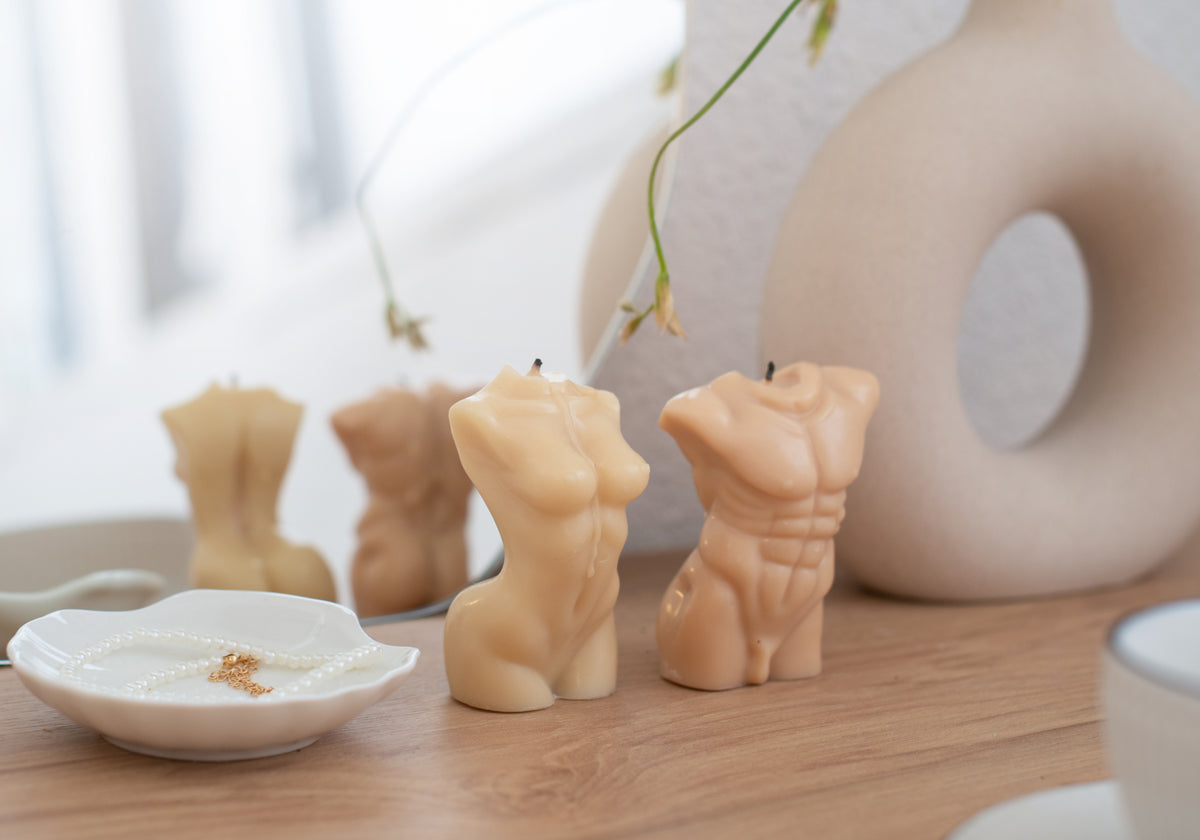 Three small torso-shaped candles in warm beige and peach tones arranged on a wooden surface, with a white dish and a blurred decorative object in the background.