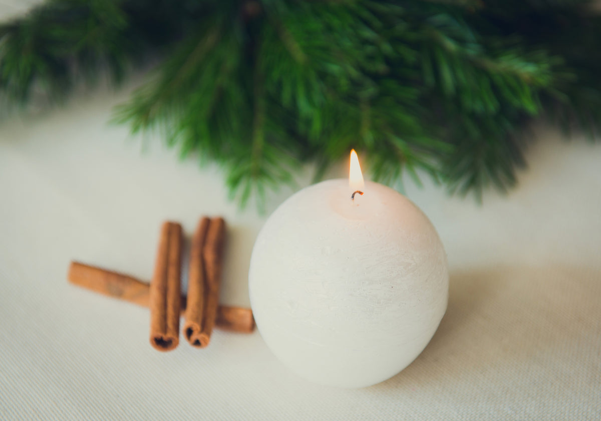 Lit white round candle styled with cinnamon sticks and evergreen branches for a warm Christmas setting.