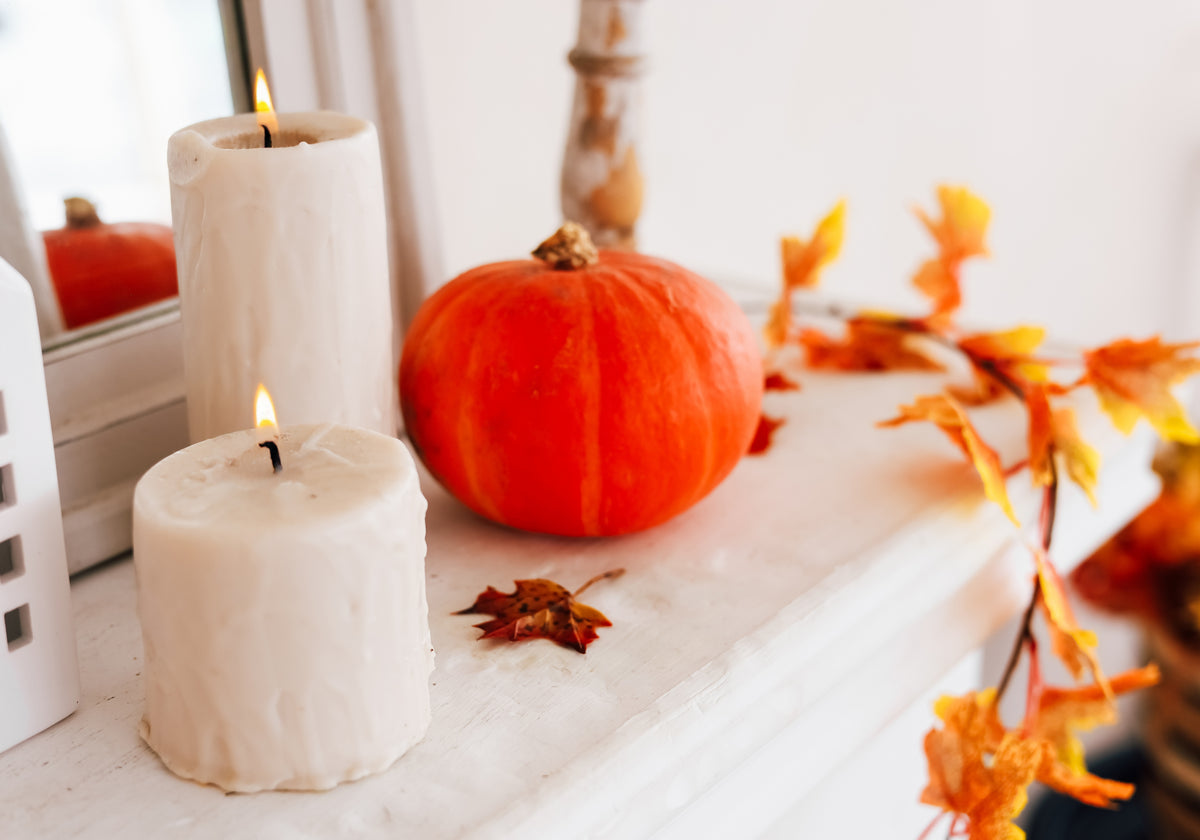 Two lit ivory pillar candles styled on a white mantel with an orange pumpkin and scattered autumn leaves for cozy fall decor.