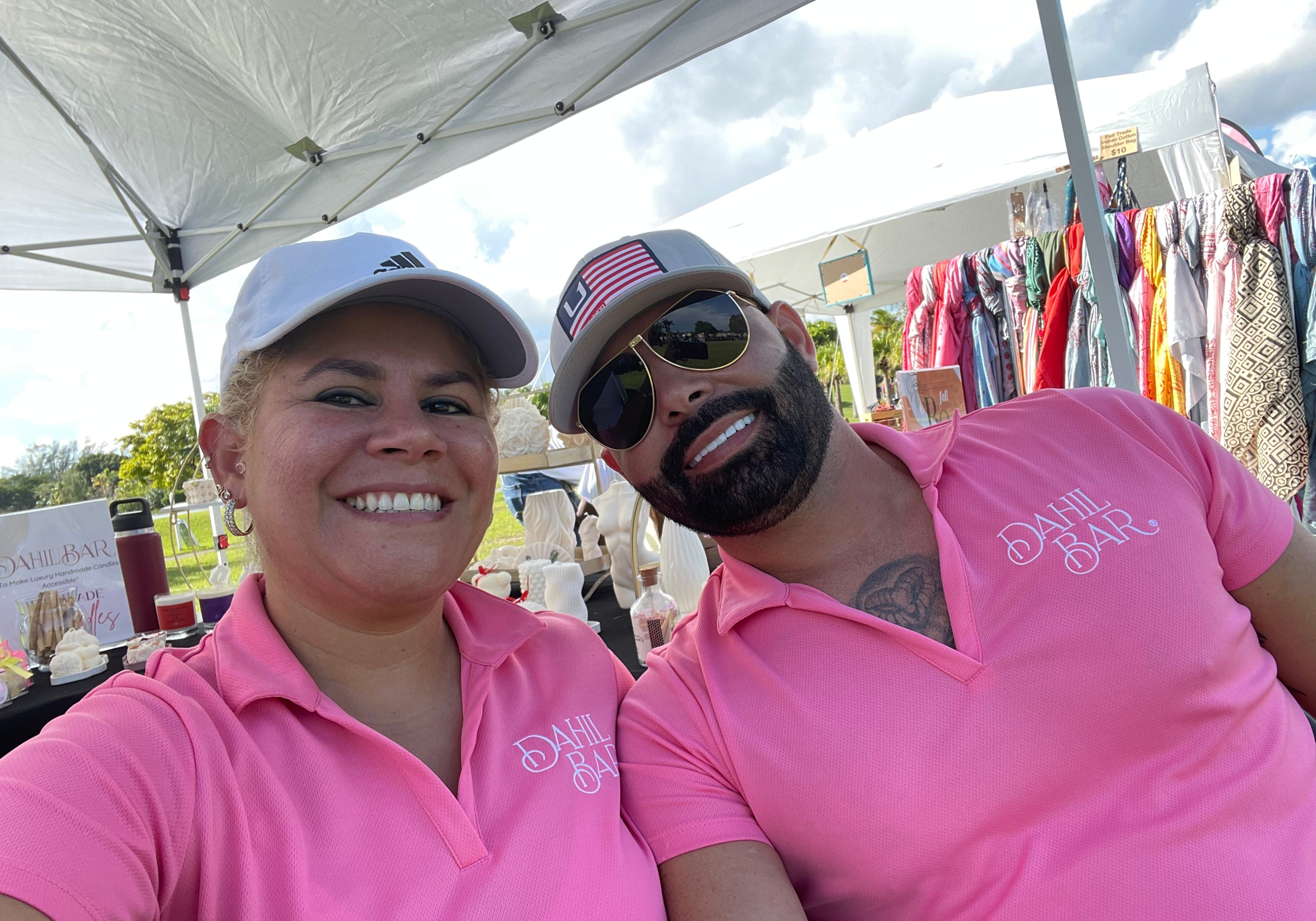 A smiling woman and a bearded man posing together outdoors under a white canopy tent. Both are wearing matching bright pink polo shirts with 'Dahil Bar' printed in white script on the left chest, along with white caps and sunglasses.