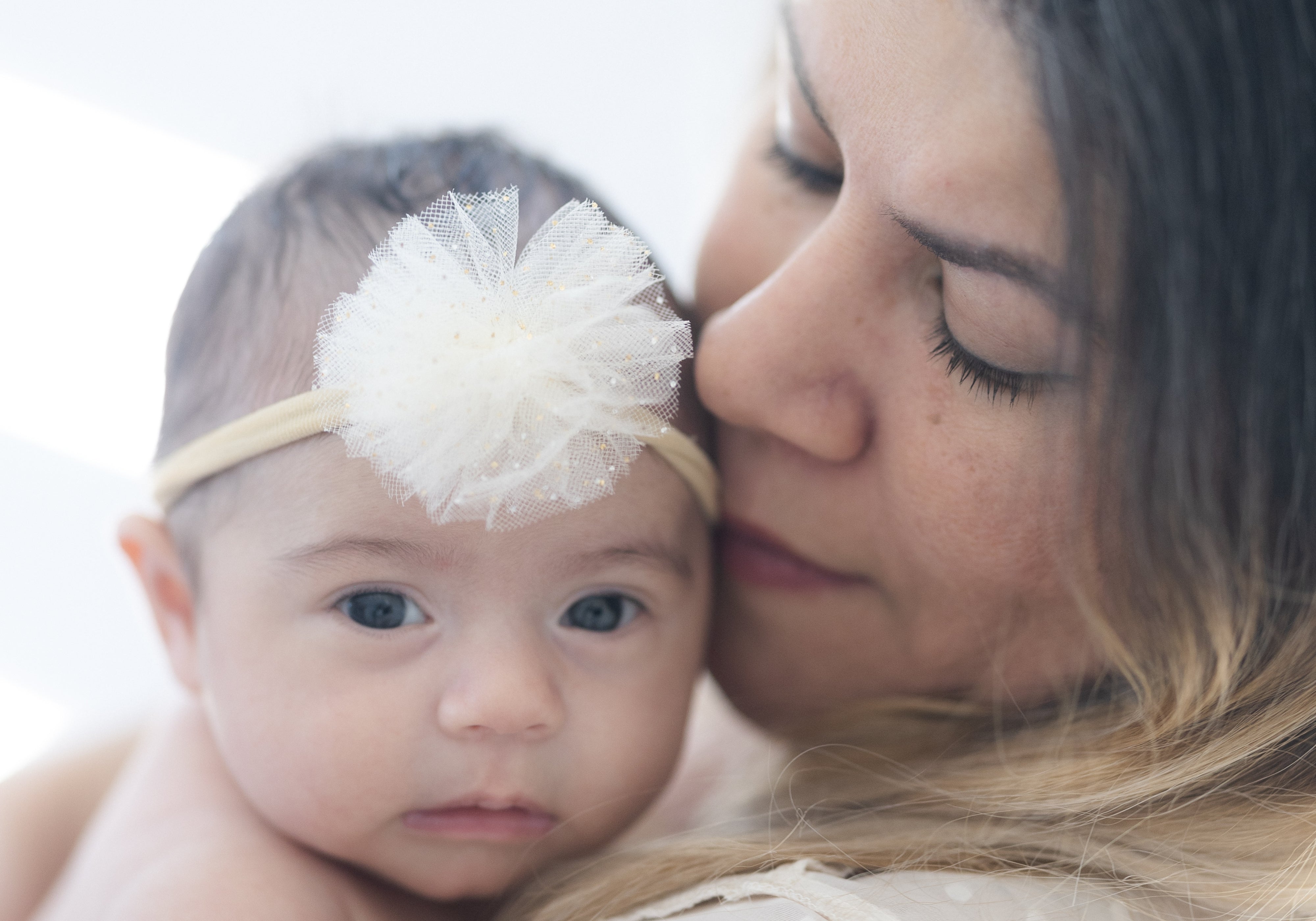 Mother kissing her baby girl wearing a cream floral headband in a soft, intimate portrait.