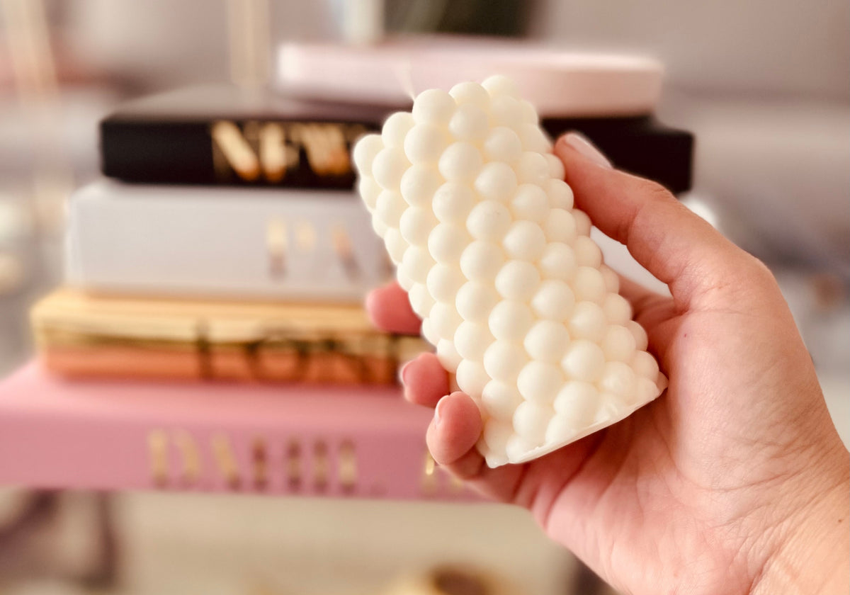 Hand holding a white bubble sculptural candle in front of stacked books, styled in a warm neutral home setting.
