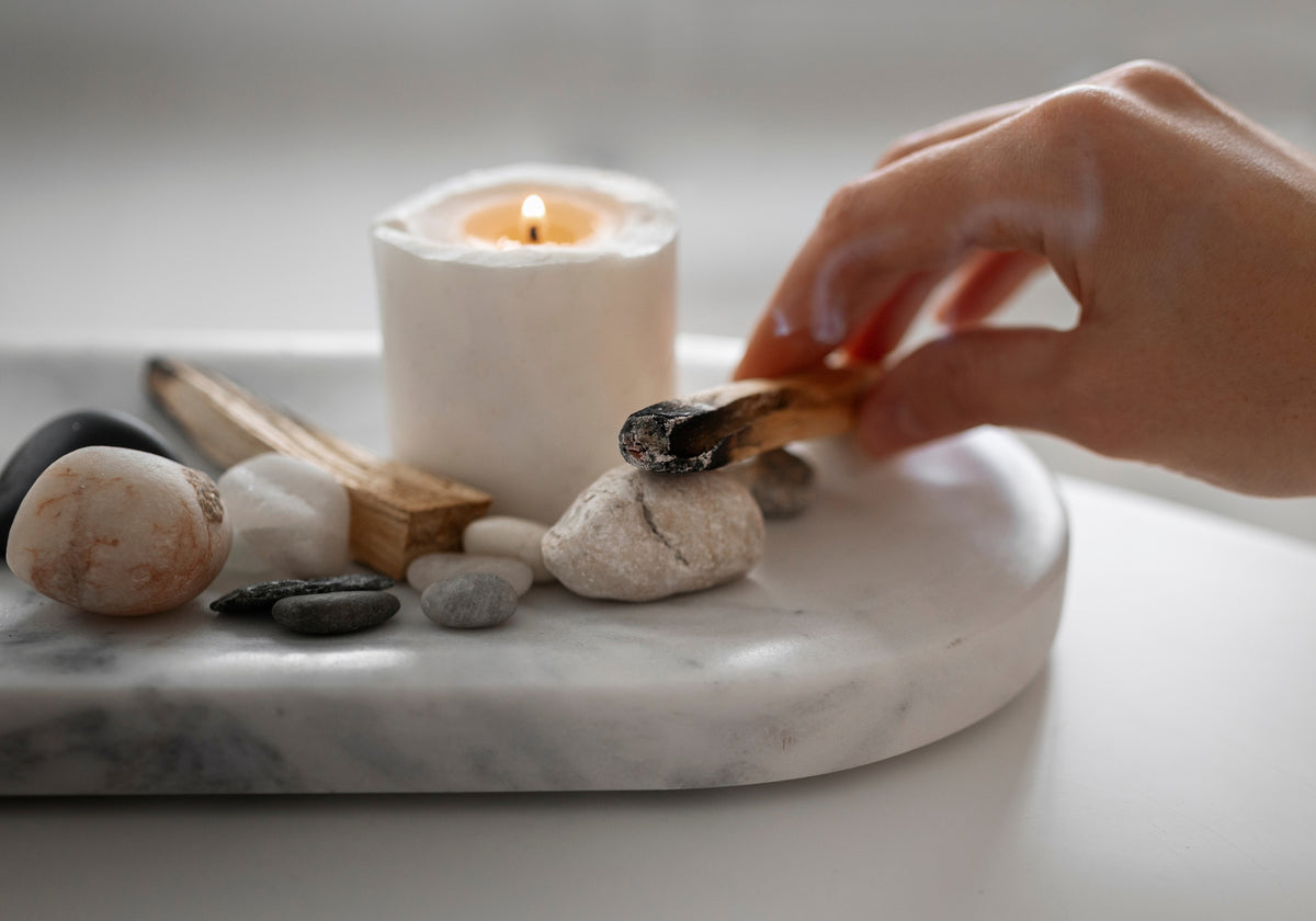 Lit white pillar candle beside natural stones and a smoldering wood stick, creating a warm, sophisticated holiday atmosphere.