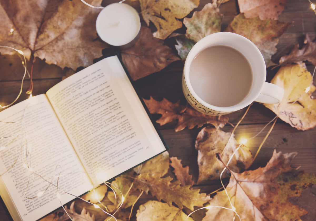 Cozy fall scene with a warm drink, open book, candle, autumn leaves, and string lights on a wooden surface.