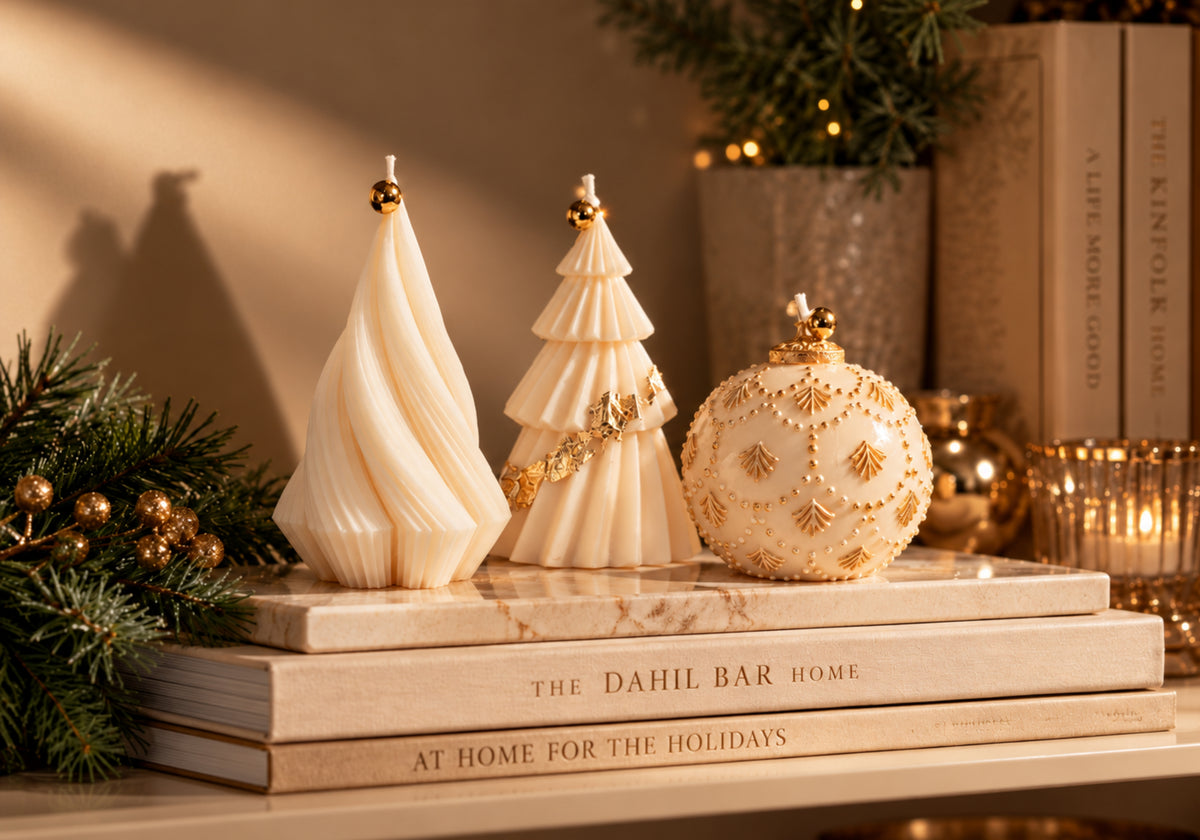 Three ivory sculptural Christmas candles with gold accents styled on stacked Dahil Bar books with evergreen branches and warm holiday lights.