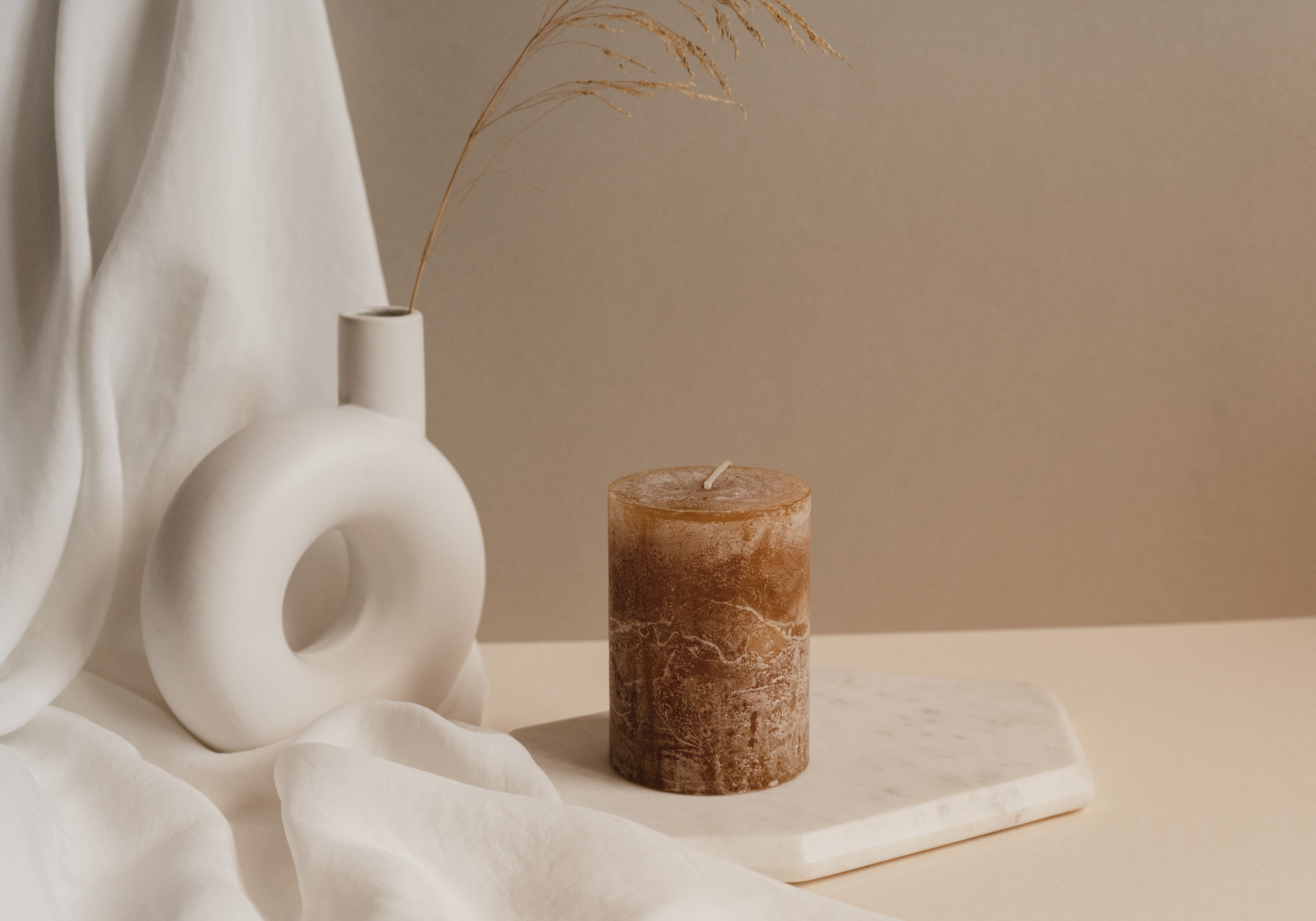 Brown pillar candle on a marble tray beside a sculptural white vase and dried grass in a warm, minimalist fall setting.