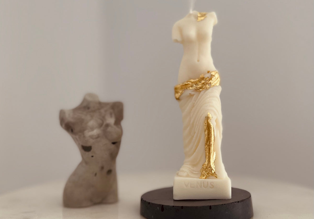 A white Venus statue candle with gold leaf accents on a black round base, displayed next to a blurred gray sculpture on a white surface against a light gray background.