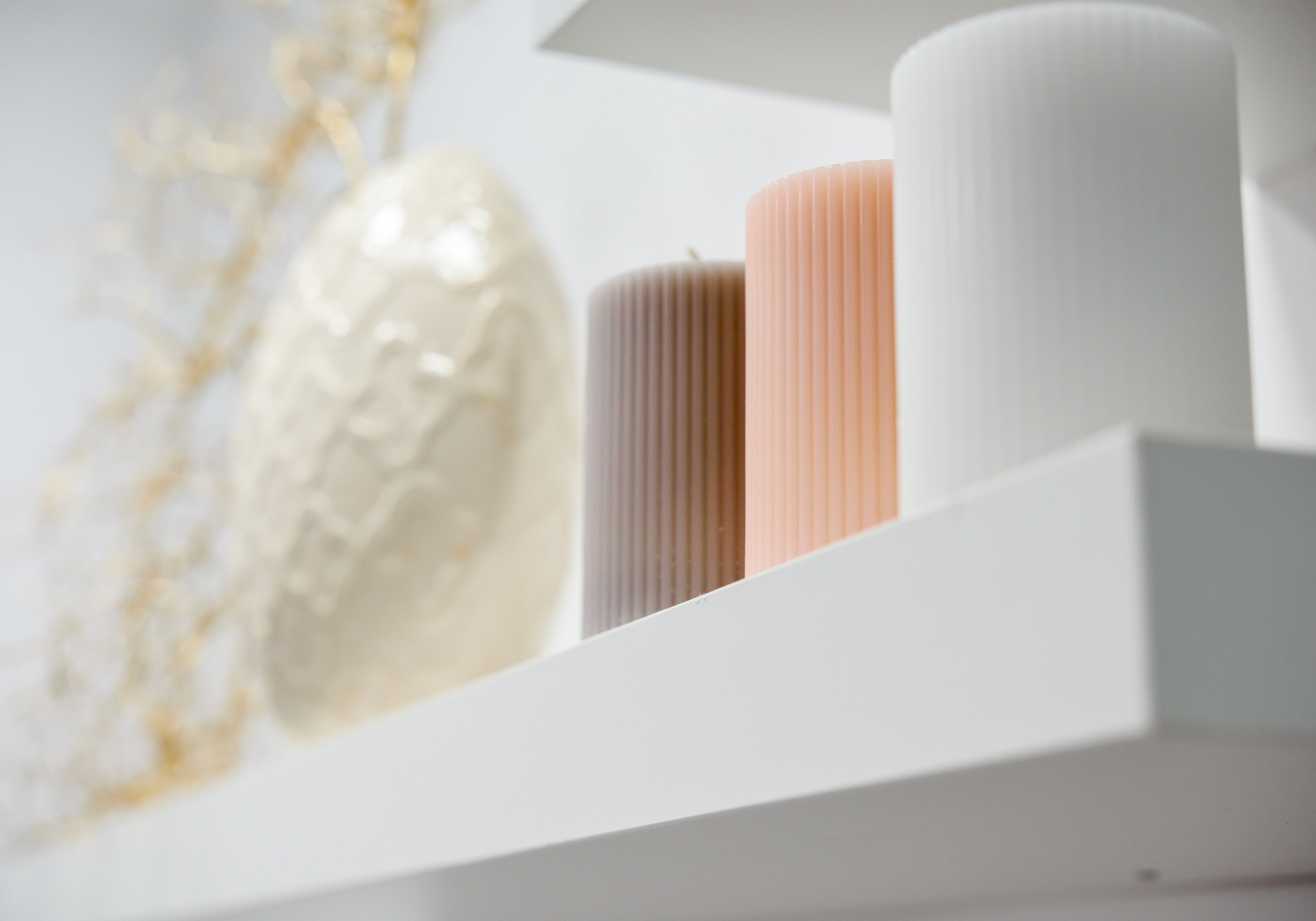 Three ribbed pillar candles in white, peach, and taupe arranged on a white shelf next to a textured cream vase and dried gold branches.