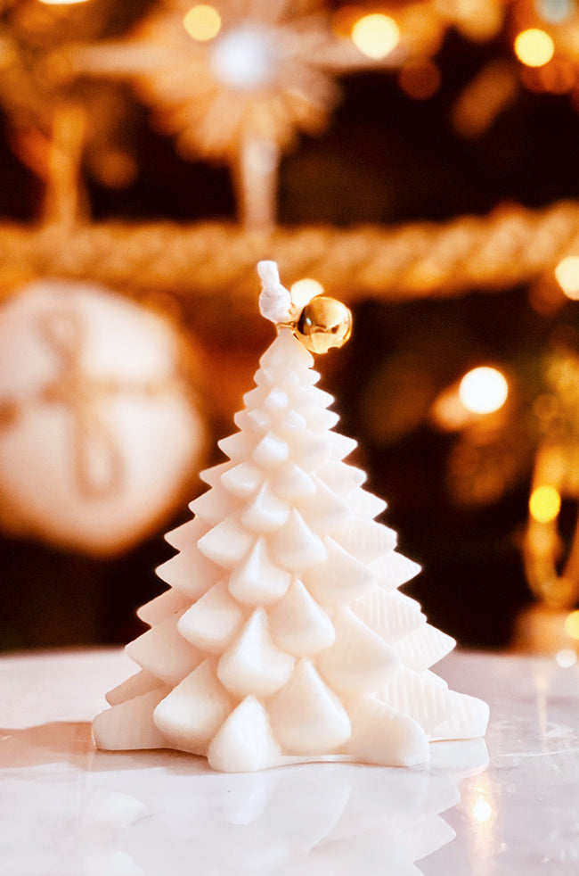 Fiery Tree Small Candle: Small ivory Christmas tree-shaped candle topped with a gold bell, styled with warm holiday lights in the background.