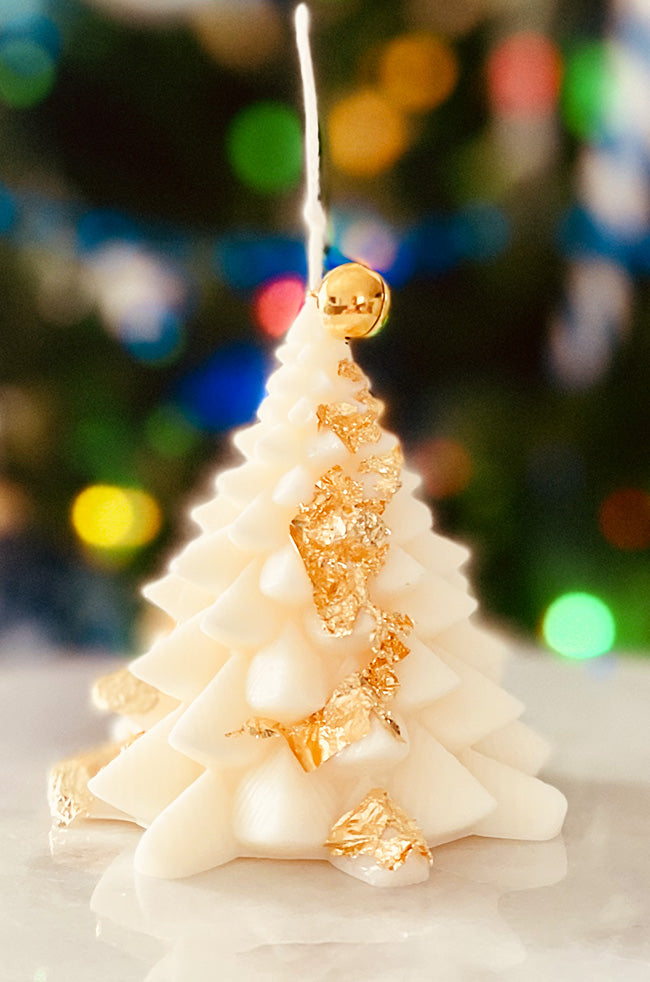 Fiery Tree Small Candle: Small ivory Christmas tree candle with gold leaf/flacks accents and a gold bell topper, set against colorful festive lights.