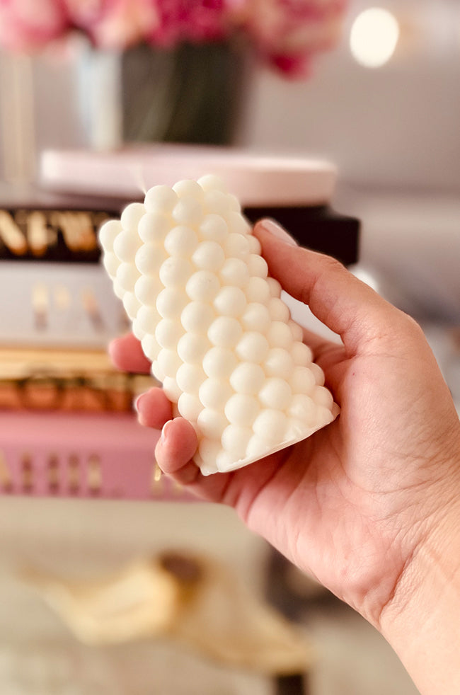 Fire Beads Candle: Hand holding an ivory sculptural candle with a stacked bead design, set against a blurred background of books and soft decor.