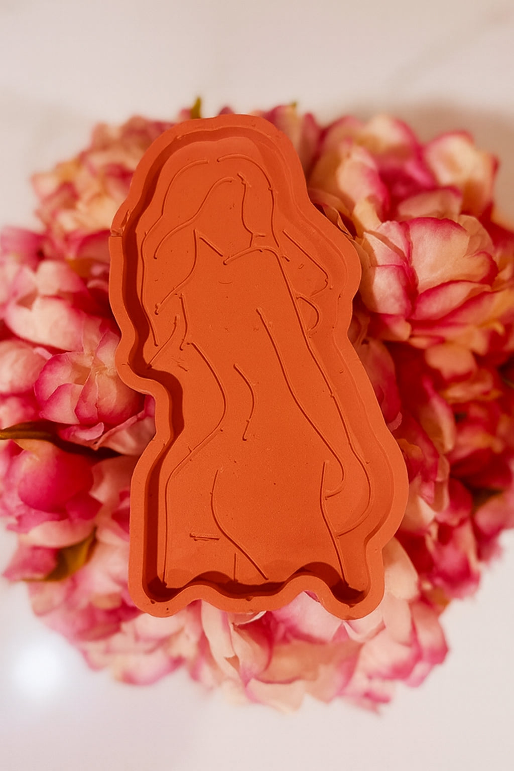 Goddess Silhouette Tray Terracota: Terracota tray with an embossed feminine silhouette design, displayed on a bed of pink flowers.