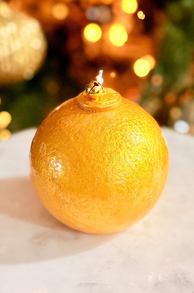 Golden Luxe Ornament Candle: A round gold ornament-style candle with textured finish and a bell accent, photographed on a marble surface with festive warm bokeh lighting.