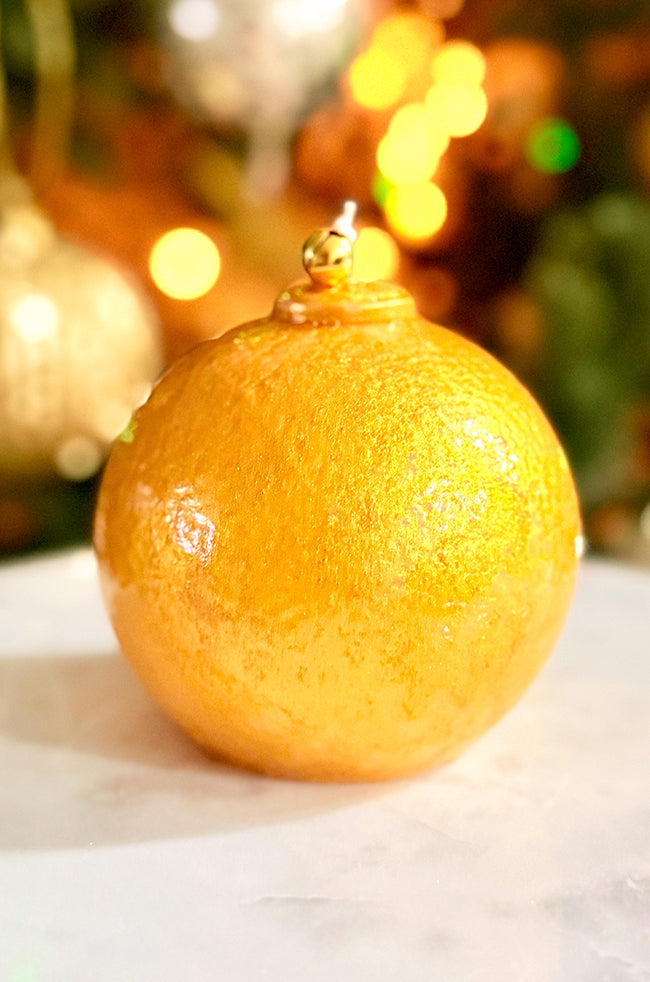 Golden Luxe Ornament Candle: Luxury gold ornament candle with a shiny hand-painted finish and a gold bell, set in front of blurred holiday décor.