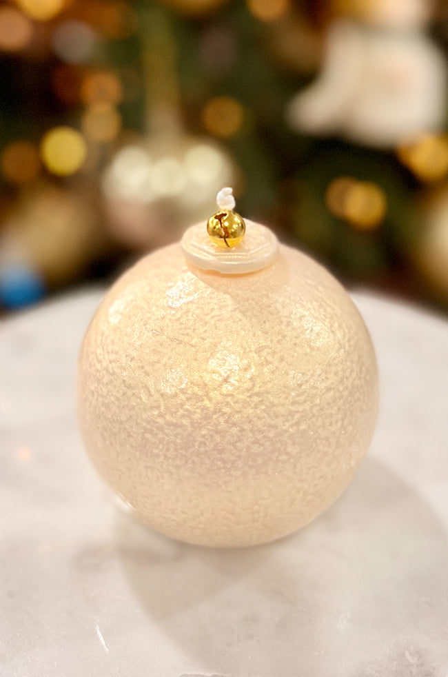 Golden Luxe Ornament:Ivory pearl ornament candle with a delicate shimmer, smooth rounded shape, and gold bell topper, photographed with festive bokeh lighting.