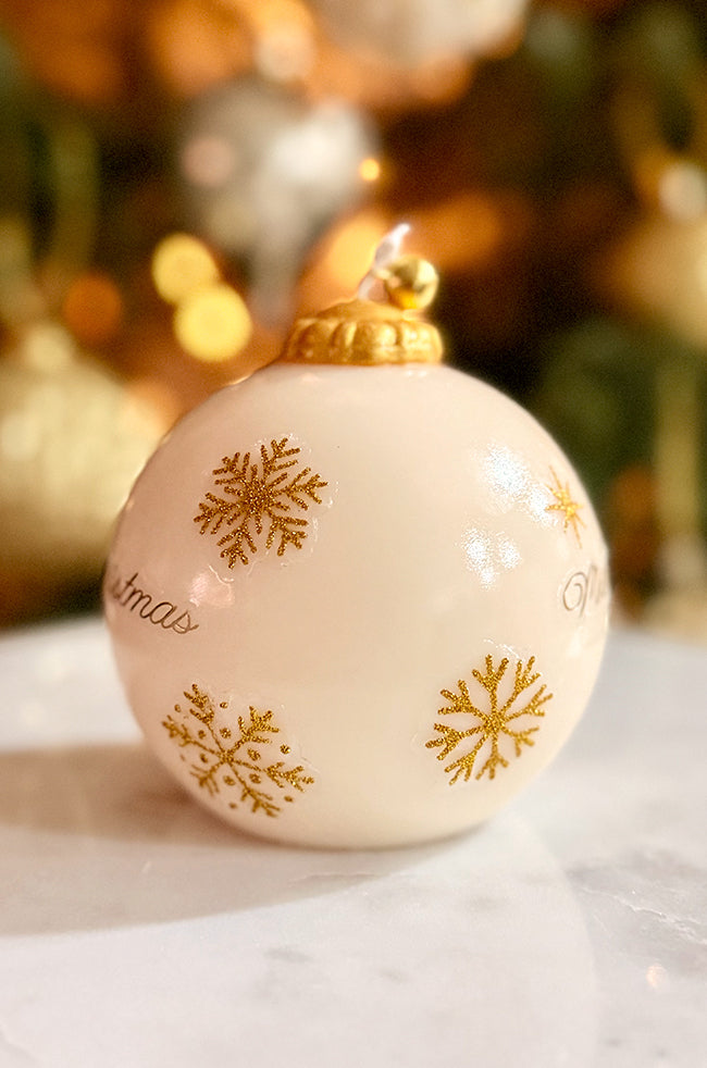 Golden Merry Ornament Candle: Close-up of an ivory ornament-shaped candle with gold snowflake accents, a polished gold cap, and bell topper, displayed in front of blurred Christmas tree lights.